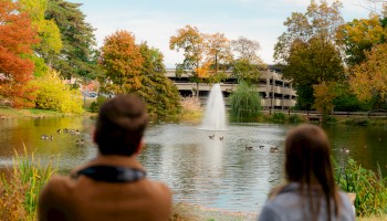 A man and woman sit by a pond with ducks, surrounded by colorful autumn trees and a fountain in the background.
