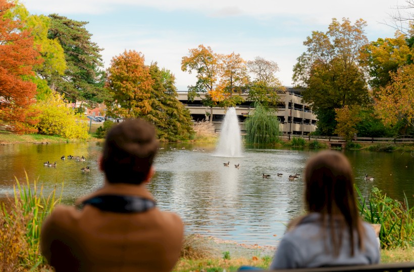 A man and woman sit by a pond with ducks, surrounded by colorful autumn trees and a fountain in the background.